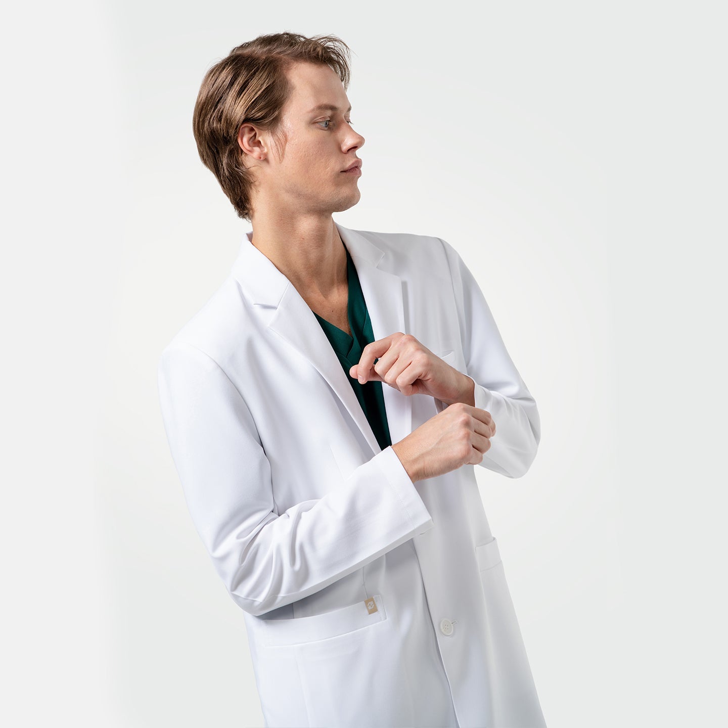 With a focused expression, a male healthcare professional in a white coat over green scrubs adjusts his sleeve