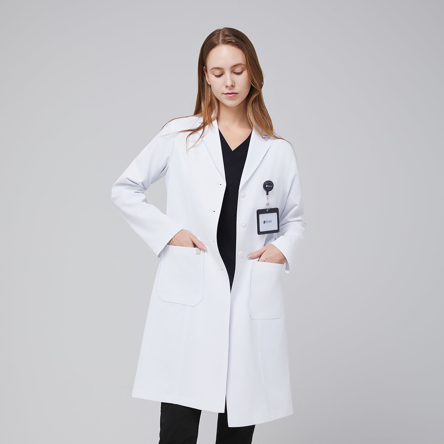 Model facing forward looking down, with a white long lab coat and black scrub top and bottom under, with her hands in her lab coat pocket,White