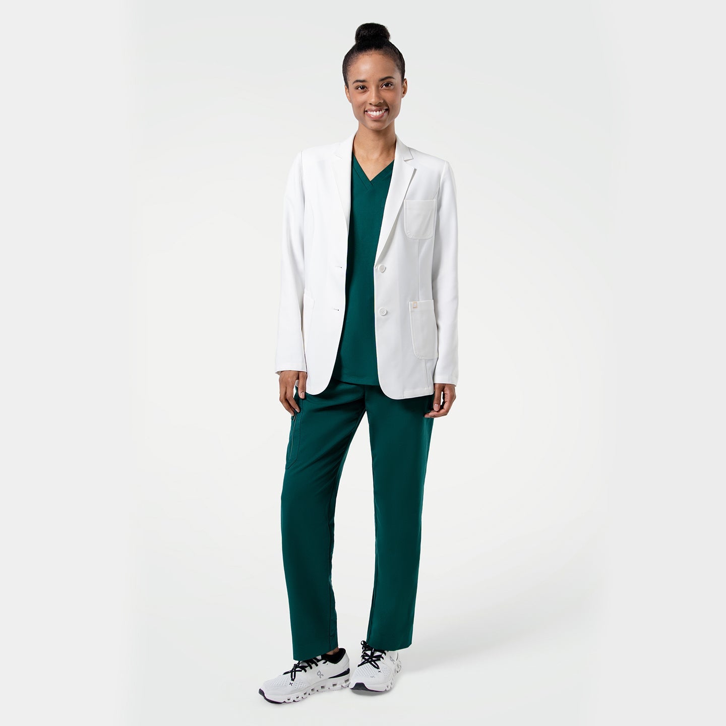 Standing pose of the model wearing the Women's LCW-101 Lab Coat over pine green scrubs, smiling with one leg slightly bent
