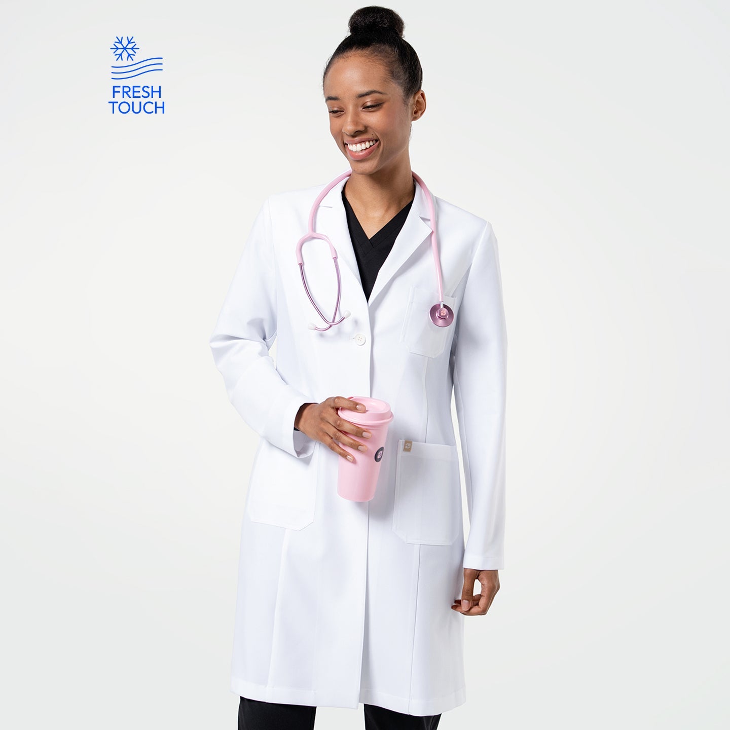 Model in the Women's LCW-102 Lab Coat in White over medical scrubs, smiling while looking slightly to the side with a pink stethoscope around her neck and holding a pink tumbler