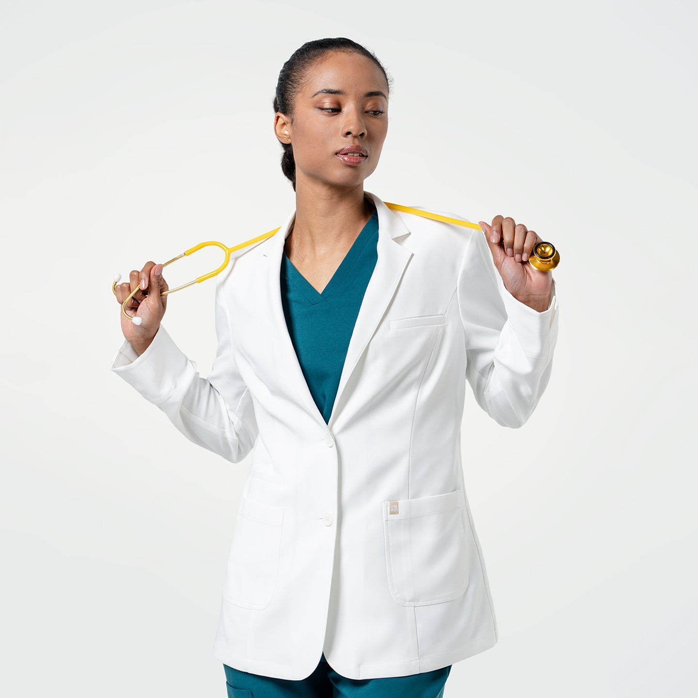 With the coat buttoned, the model wearing the Women's LCW-04 Lab Coat in White over medical scrubs holds a yellow stethoscope around her neck with both hands, upper body view