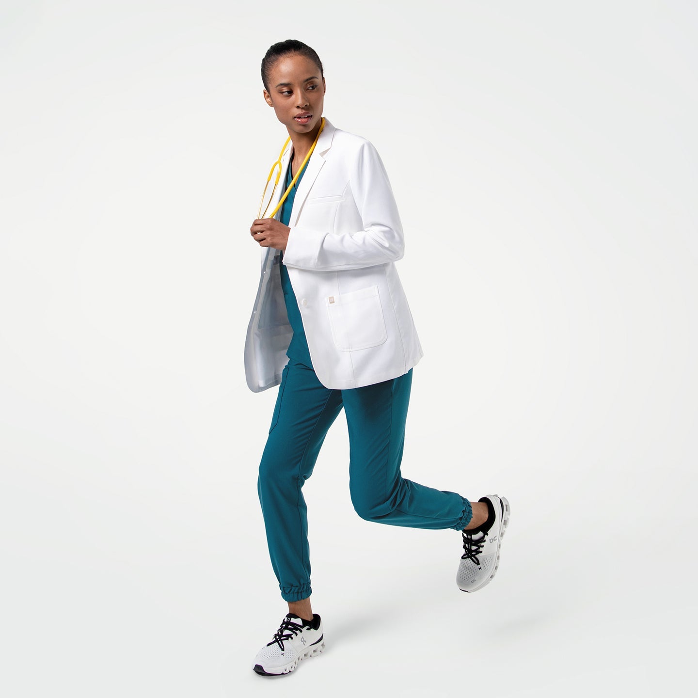 Captured in motion, the model wears the Women's LCW-04 Lab Coat in White over medical scrubs, running toward the camera with her body slightly angled and a yellow stethoscope around her neck