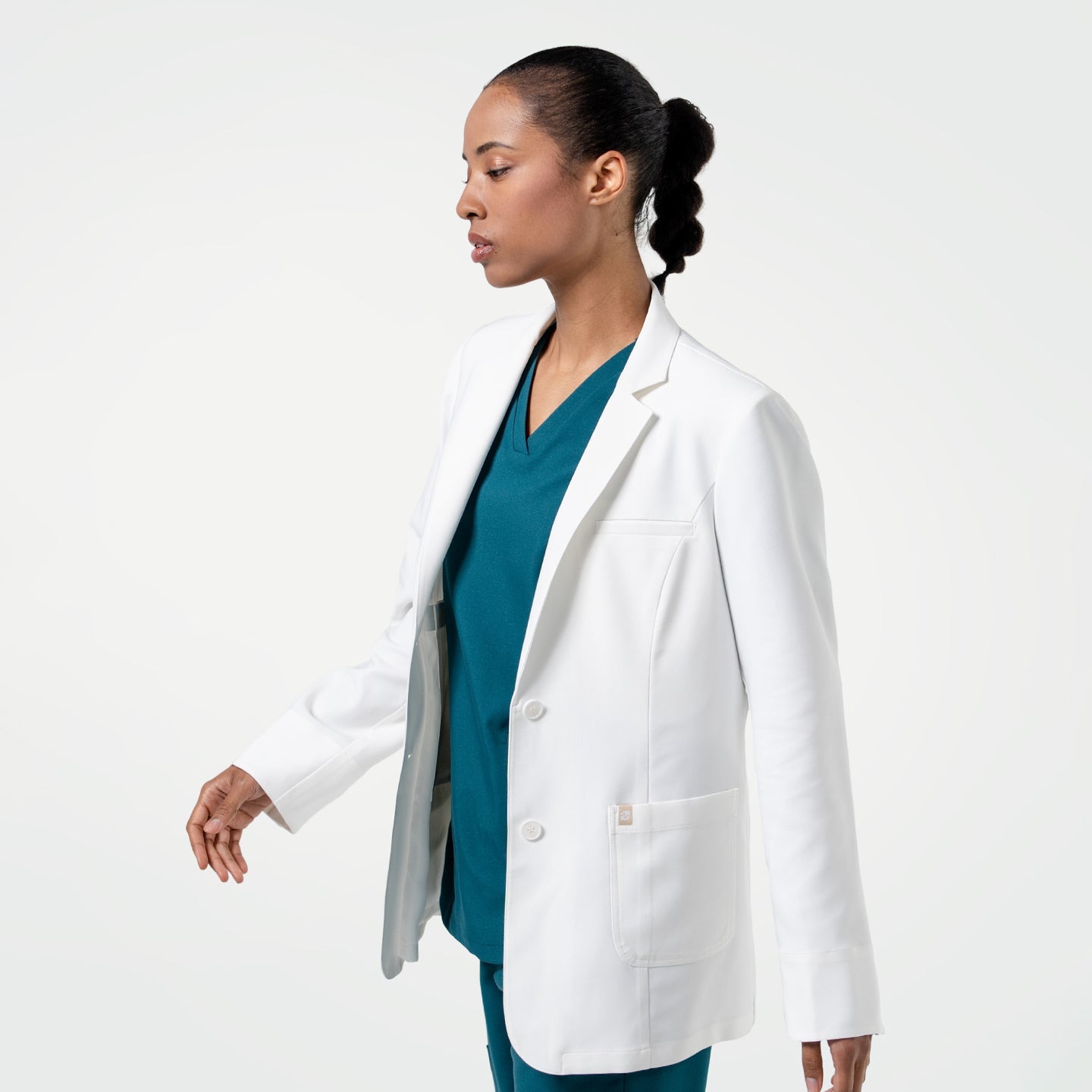 Mid-step, the model in the Women's LCW-04 Lab Coat in White over medical scrubs is captured from the side, upper body view only