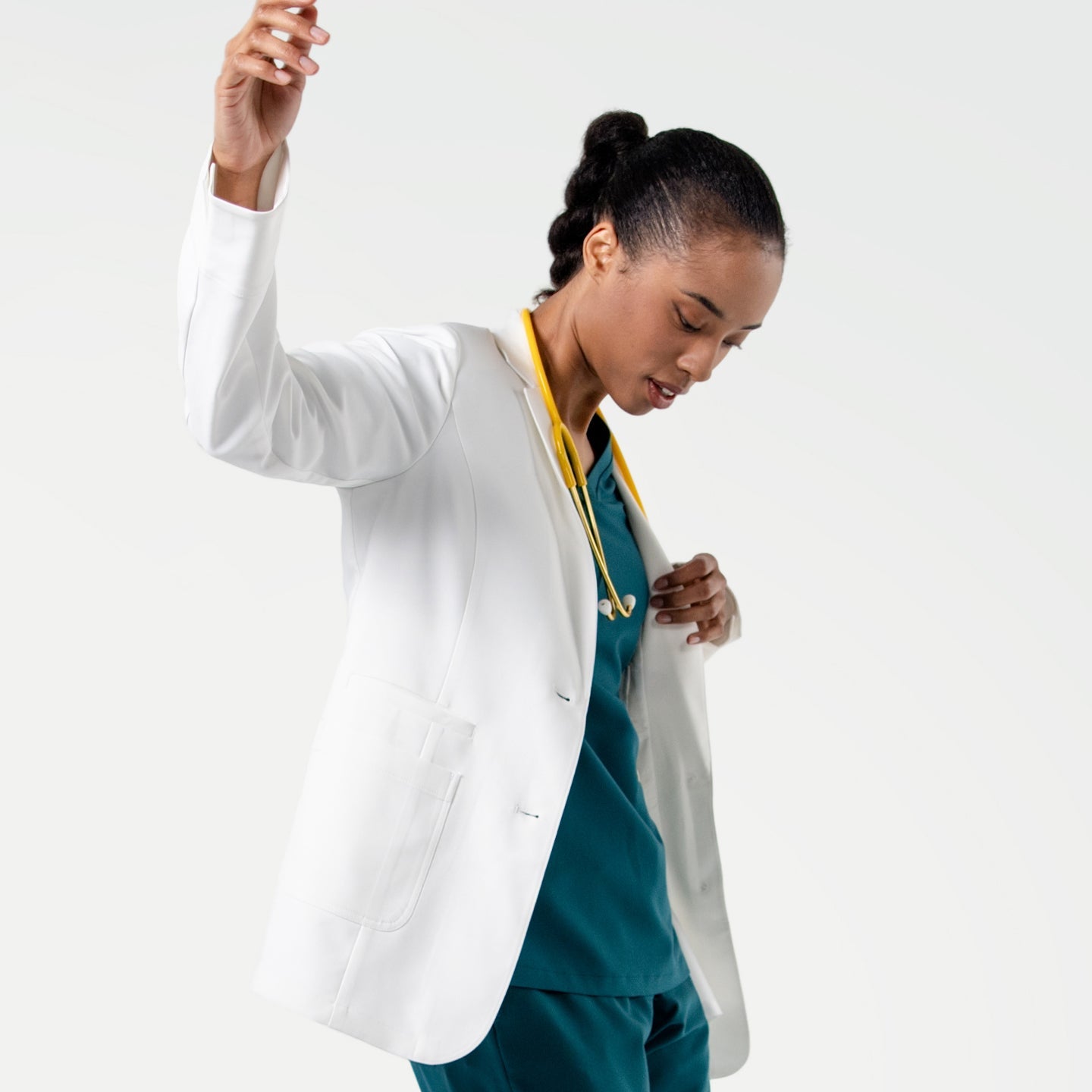 In a dynamic pose, the model wears the Women's LCW-04 Lab Coat in White over medical scrubs, with a yellow stethoscope around her neck and one hand slightly pulling the coat aside