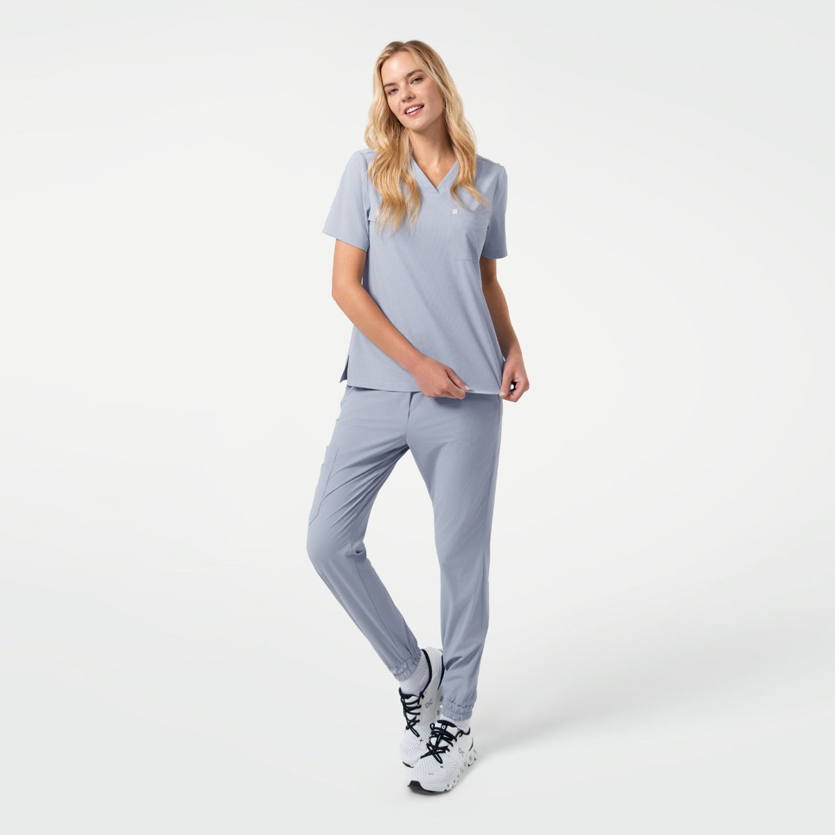 A woman walking while wearing StretchFit Jogger Scrub Pants in Cool Gray with a matching V-neck scrub top and white sneakers.