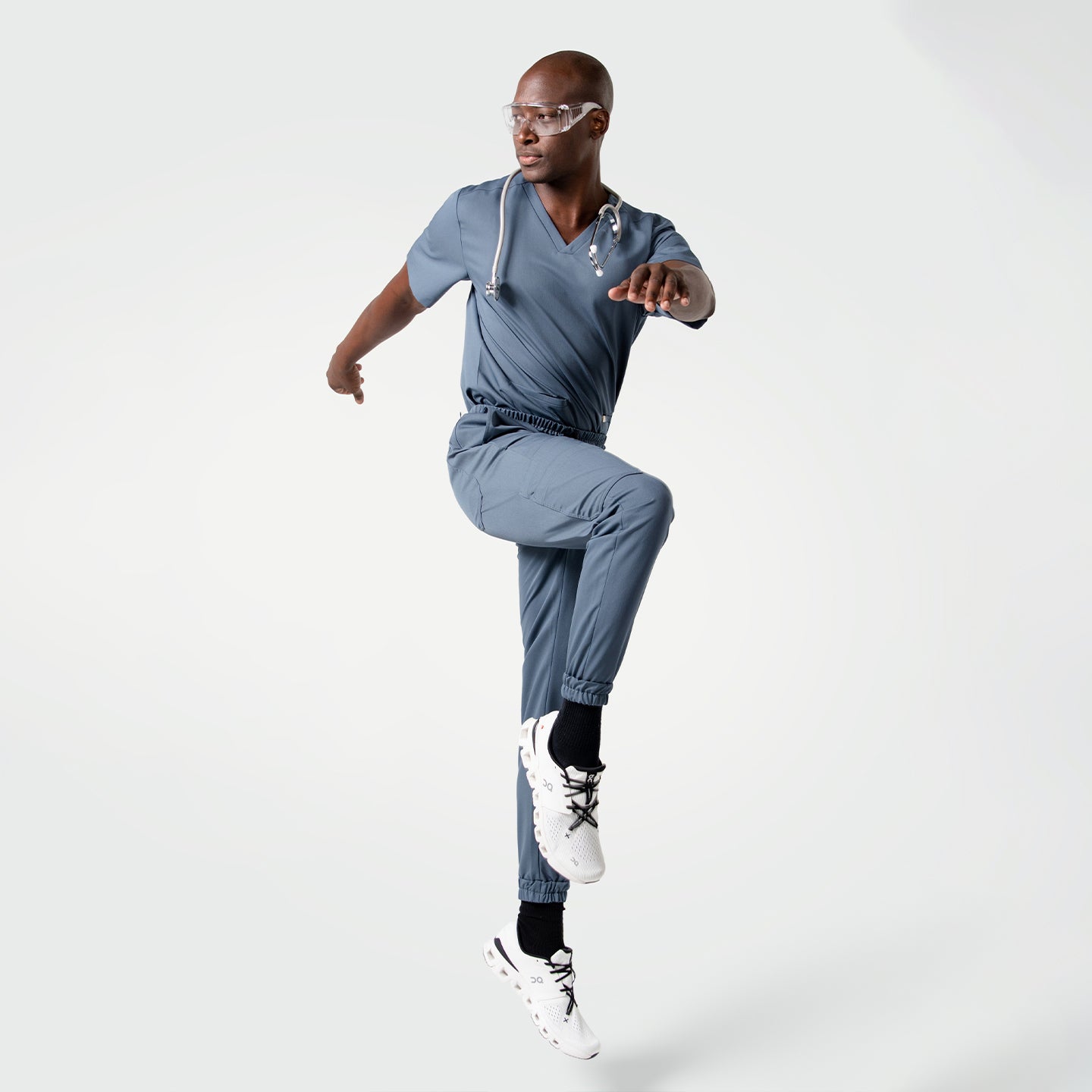 Model performing a dynamic jump pose while wearing the Smoky Blue scrub set with a stethoscope around the neck.