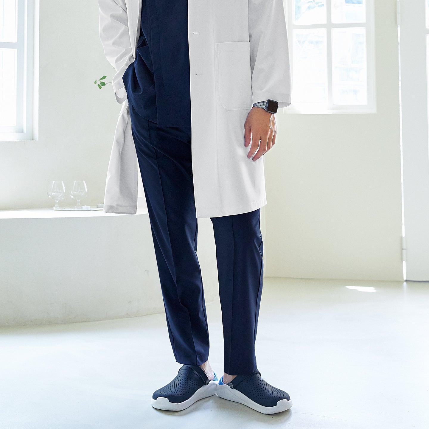 Close-up of man wearing a long lab coat over a front zipper scrub top with chest and side pockets, paired with straight-leg scrub pants, standing indoors,Eco Navy