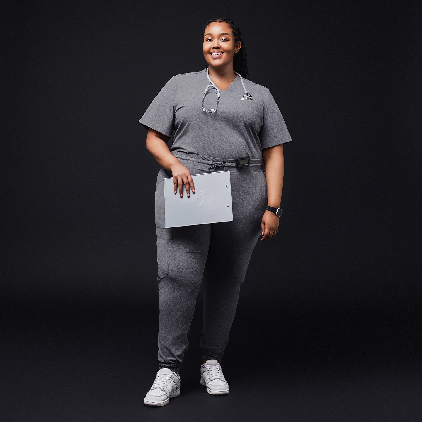 Smiling woman in ash gray scrub set with jogger pants, stethoscope, and white sneakers, holding a clipboard on a black background,Ash Gray