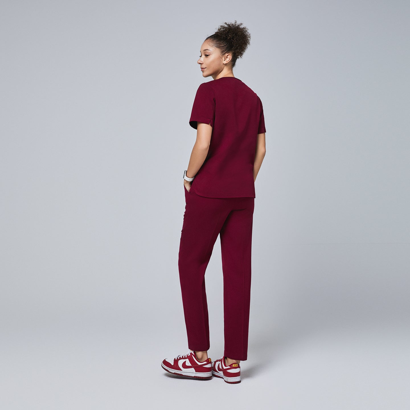 Female model wearing a burgundy scrub top with short sleeves and matching pants, shown from the back with hands in pockets, paired with burgundy and white sneakers,Burgundy