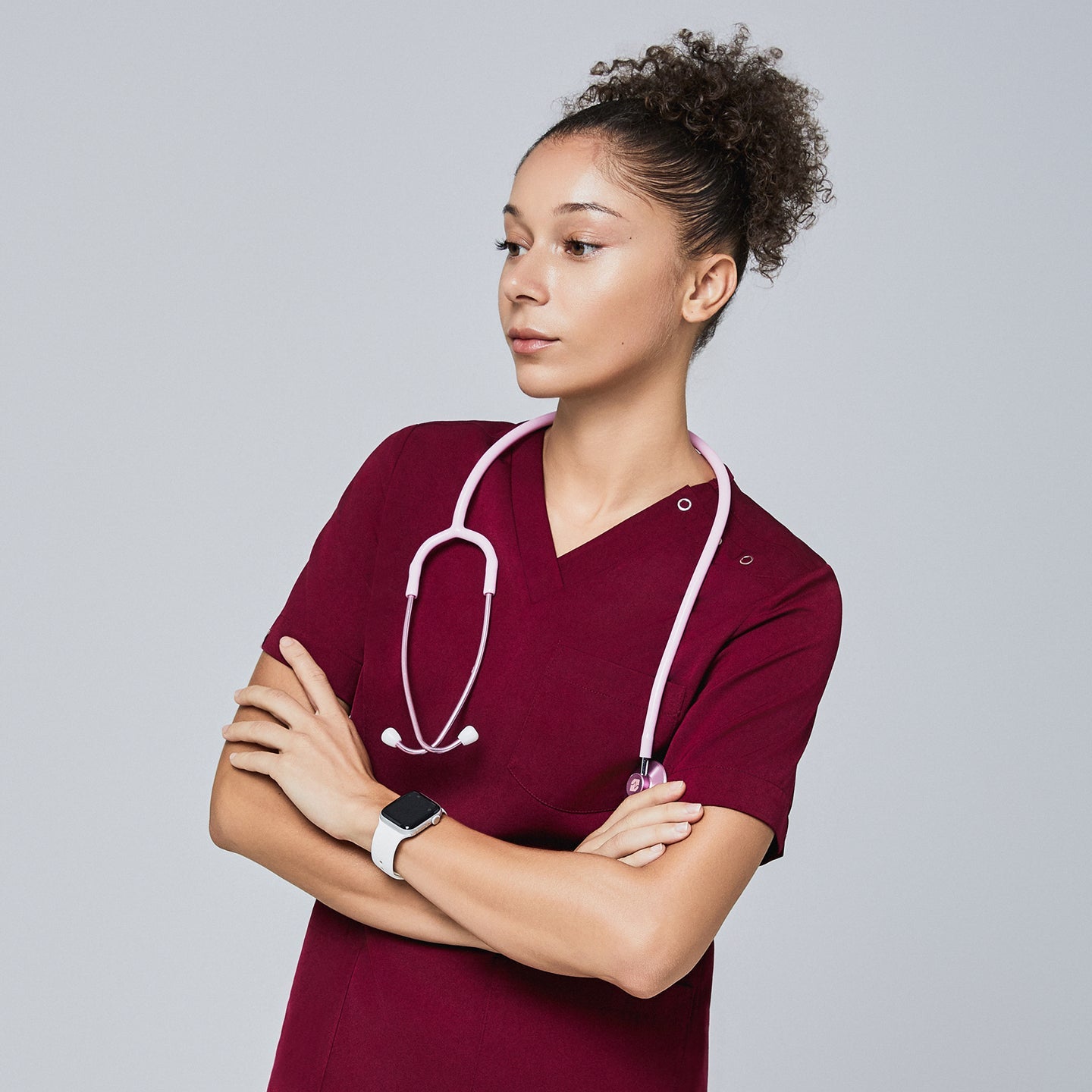 Female model wearing a burgundy V-neck scrub top with three-button detail, accessorized with a pink stethoscope around her neck and a smartwatch, standing with arms crossed and looking to the side, Burgundy