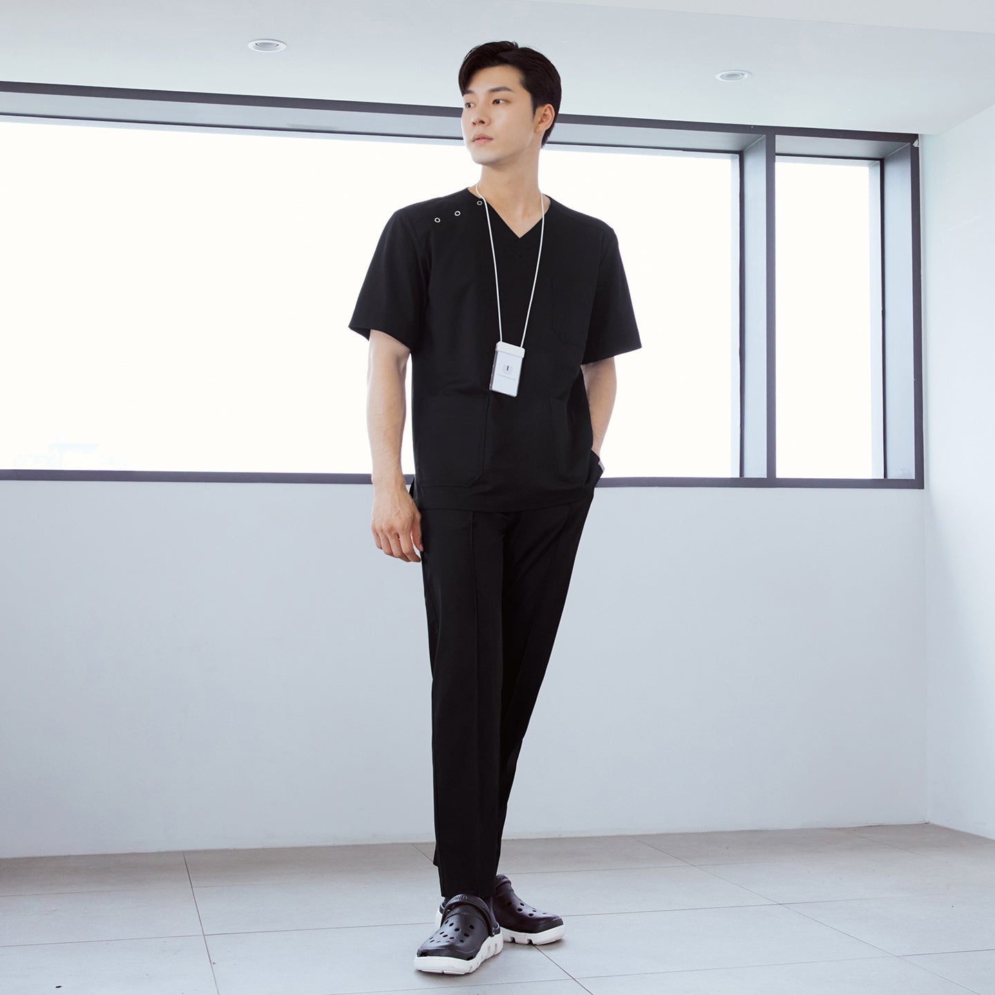  Man wearing front zipper scrub top with chest and side pockets, paired with banding scrub pants, standing indoors with hands in pockets,Eco Black
