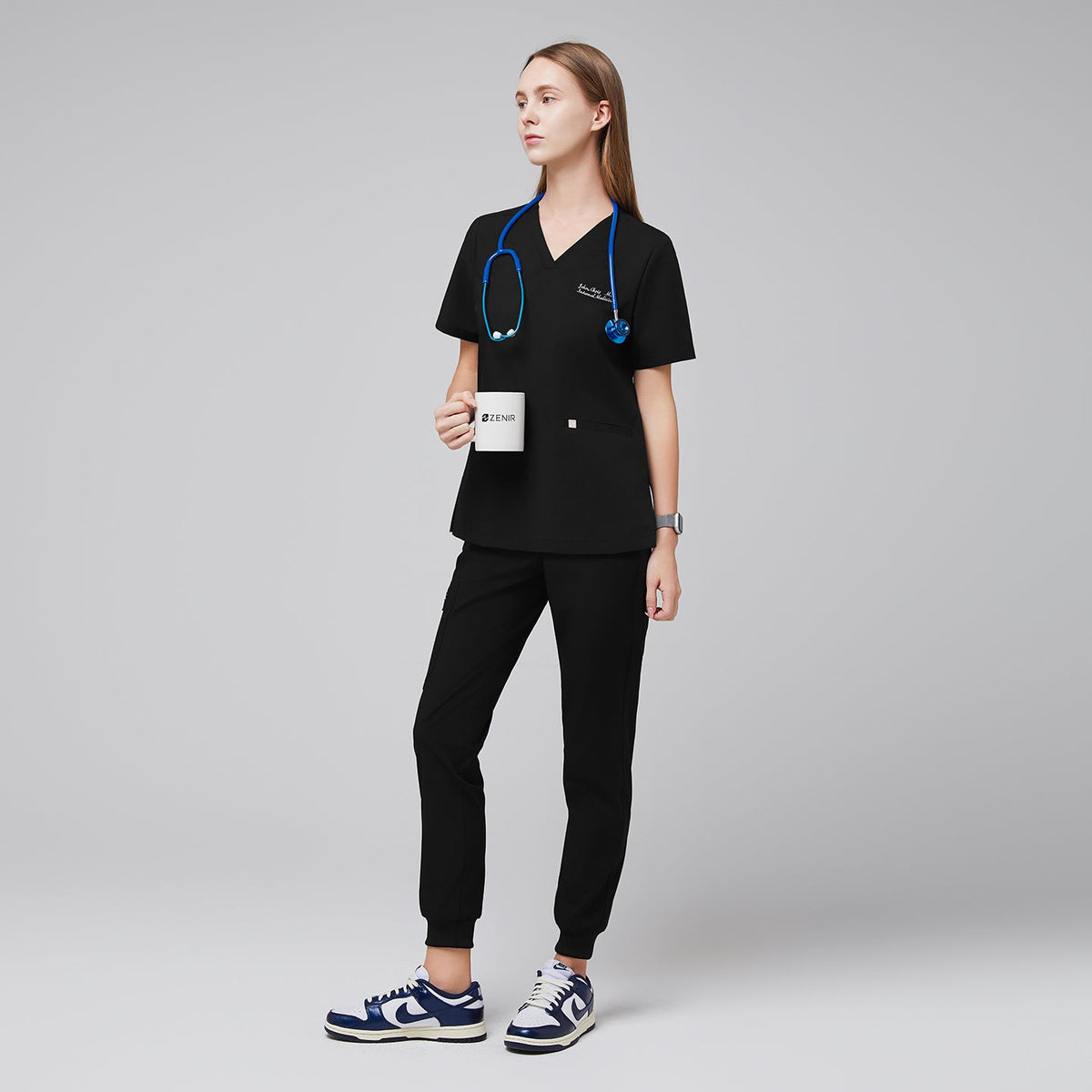 Model wearing eco black jogger scrub pants with zipper pocket and matching scrub top, styled with a blue stethoscope and navy sneakers,Eco Black