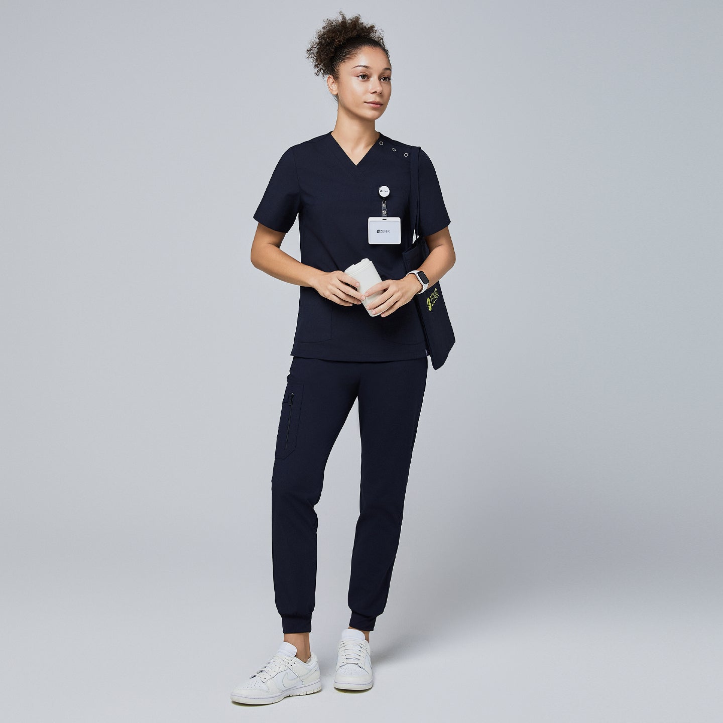 Female model wearing a navy V-neck scrub top with shoulder buttons, navy jogger scrub pants, a name badge, and a Zenir tote bag, holding a reusable cup,Eco Navy