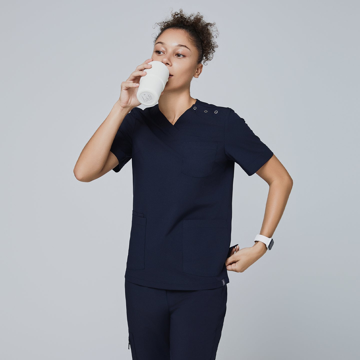 Eco navy scrub top with shoulder buttons, v-neck, and two front pockets. Model holding tumbler and placing hand in pocket for a functional look,Eco Navy