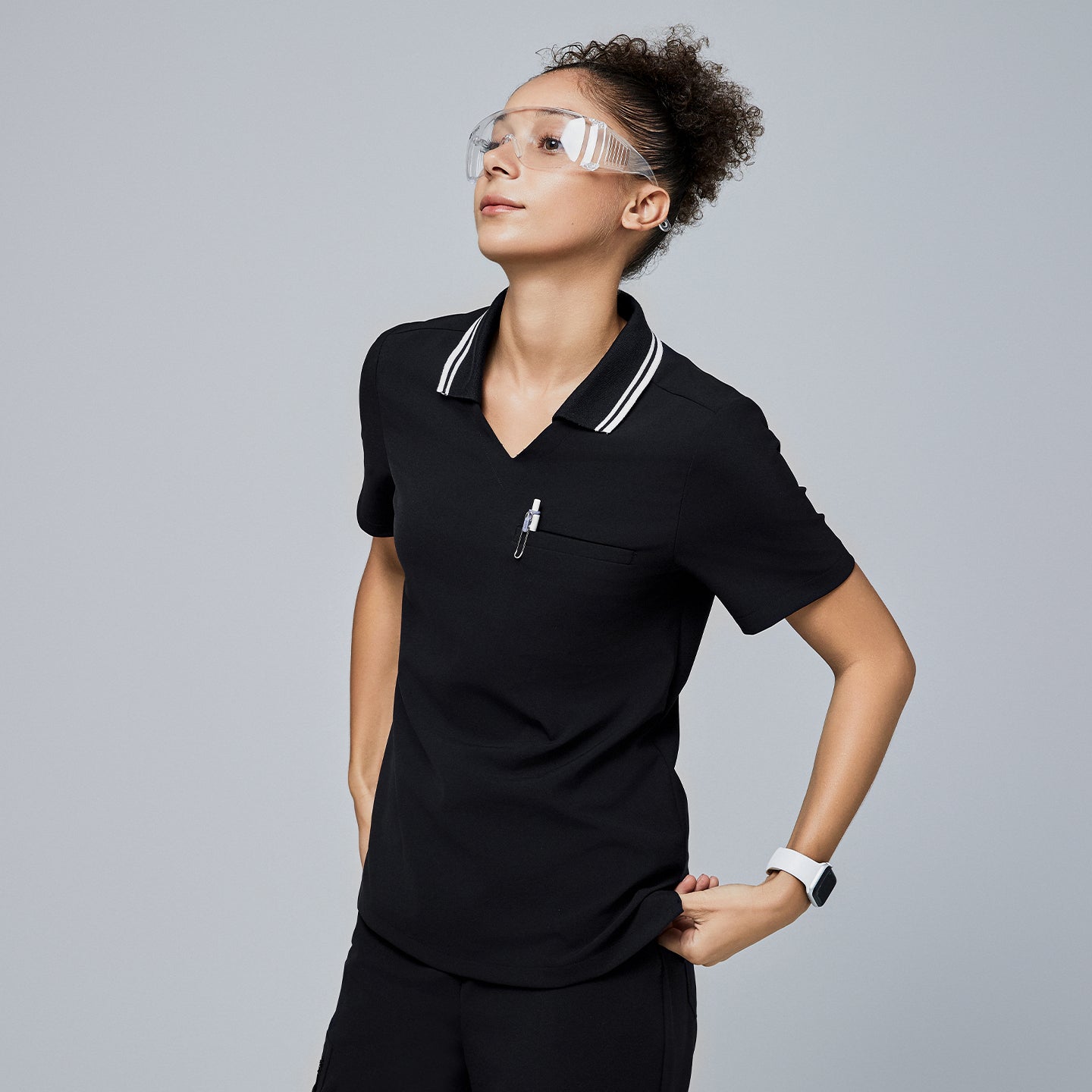 Female model wearing a jet black essential polo scrub top with a striped collar, looking upward, accessorized with safety glasses and a smartwatch,Jet Black