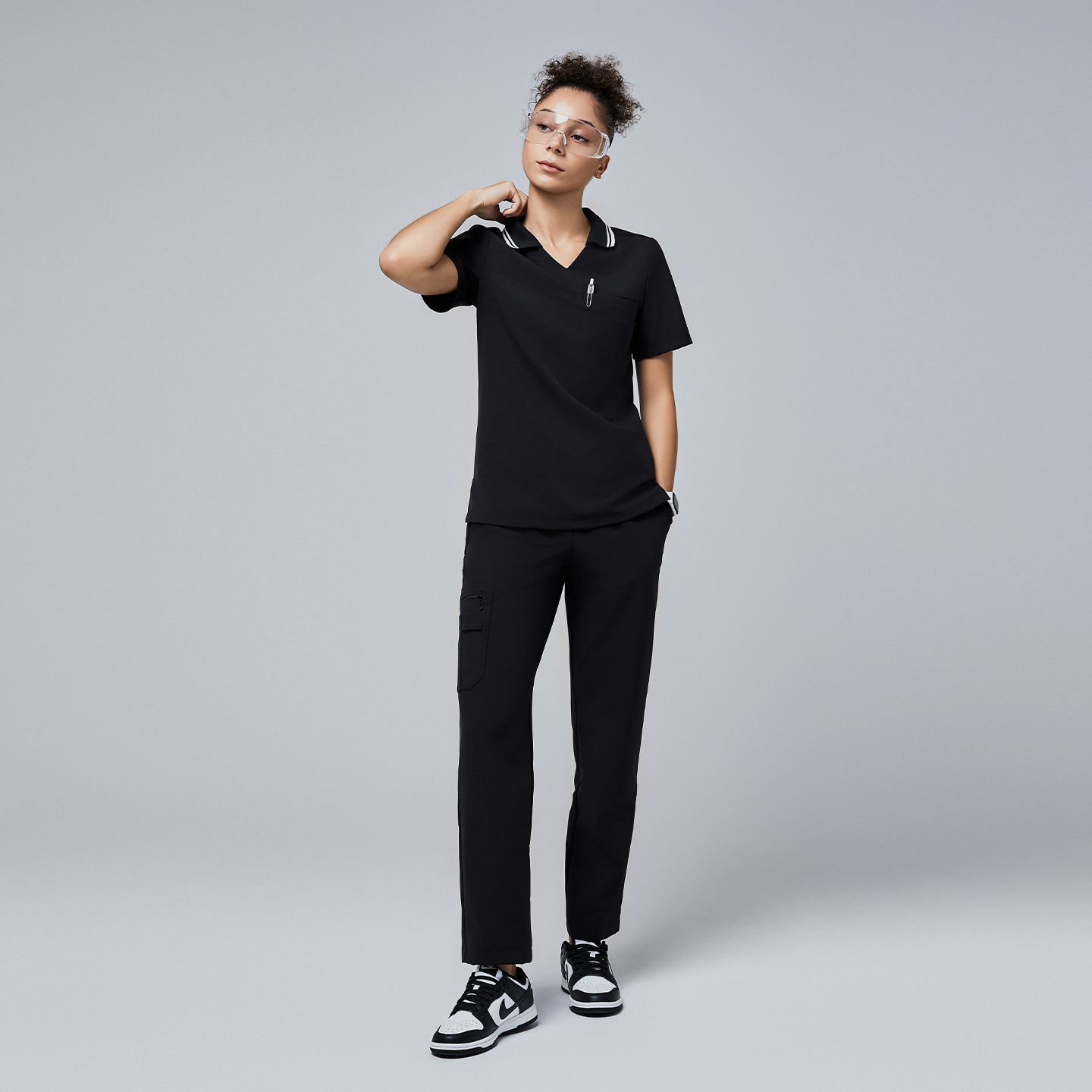 Full-body view of a woman wearing a black polo scrub top with a white striped collar, black scrub pants, and black-and-white sneakers,Jet Black