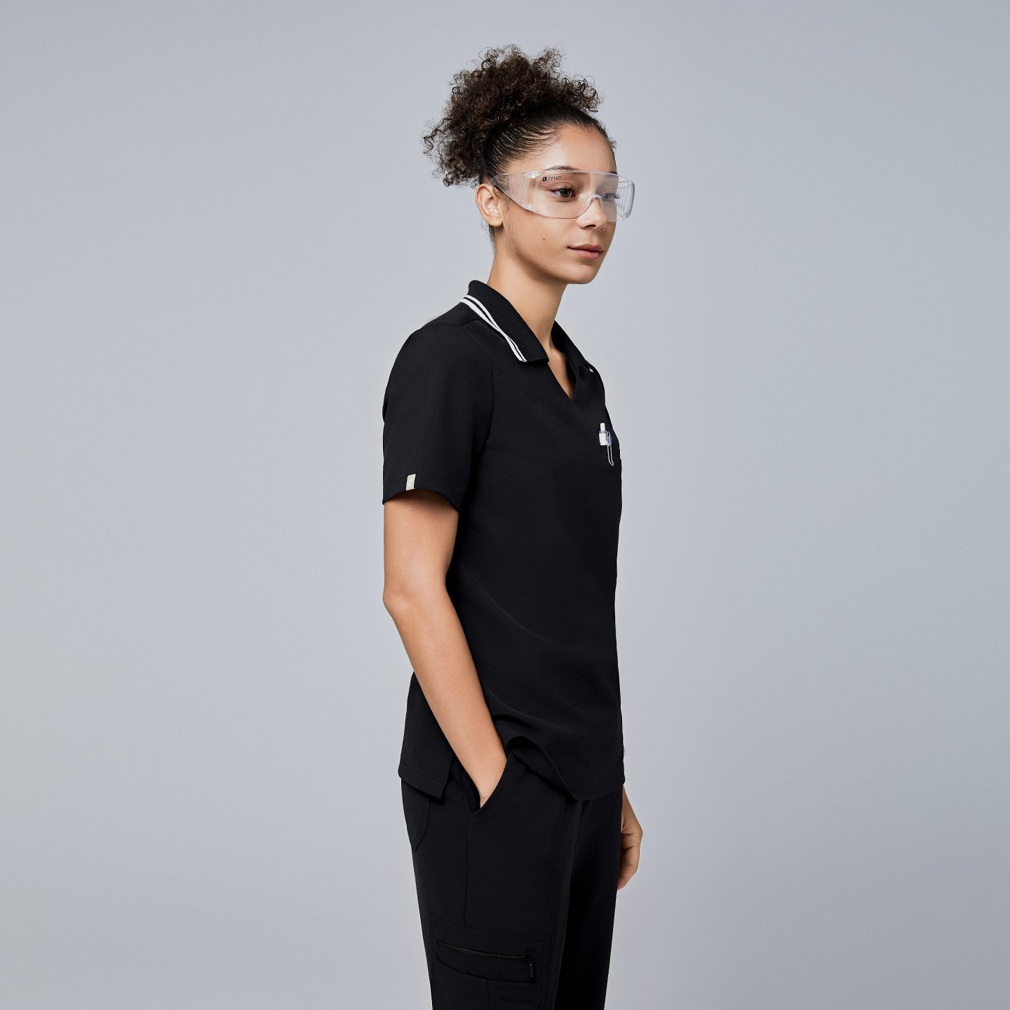 Female model wearing a jet black essential polo scrub top with a striped collar, viewed from the side, accessorized with safety glasses and her hands in her pockets,Jet Black