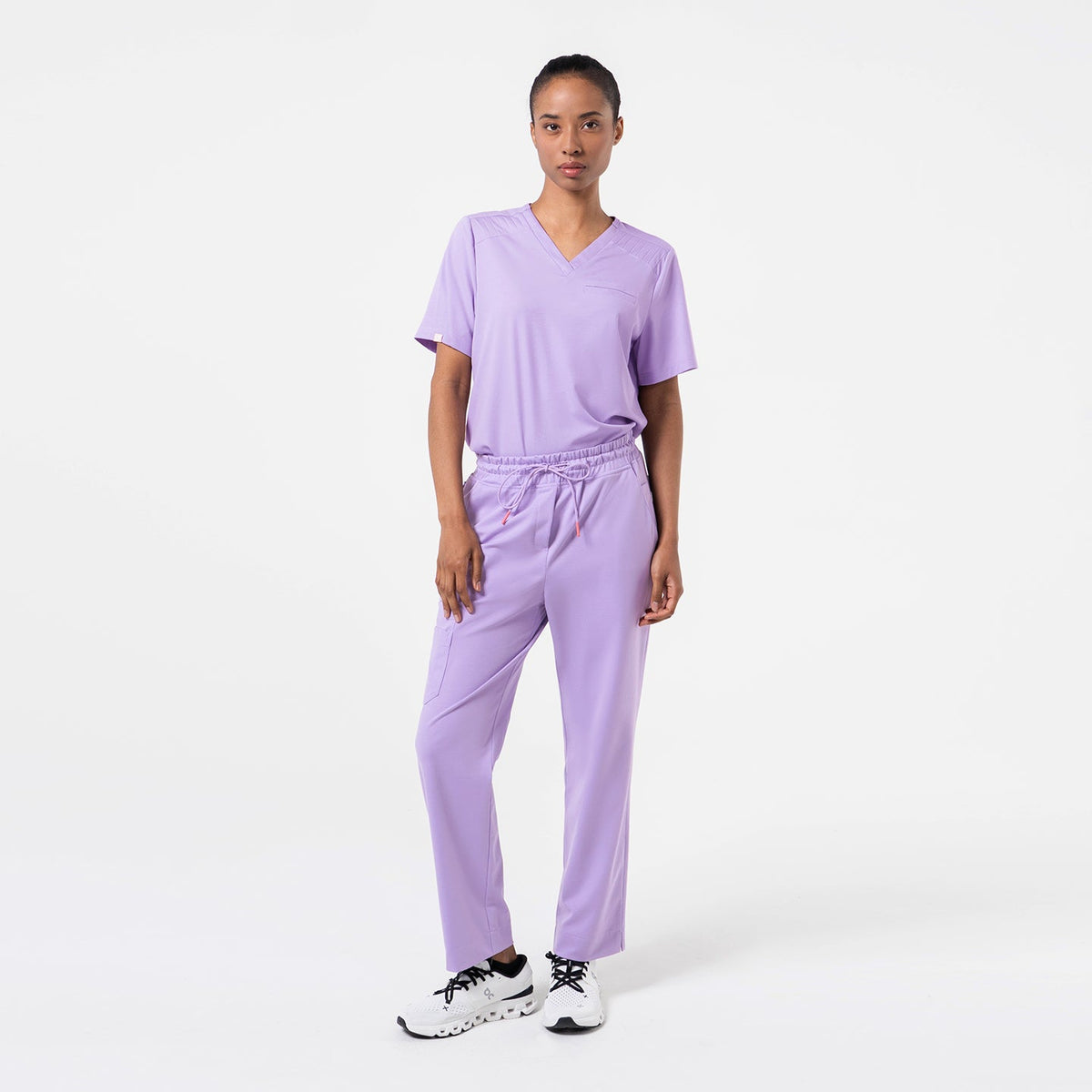 A model wears a lavender pleated-shoulder scrub top with a clean V-neck and subtle pleat detailing along the shoulders. The lightweight lavender fabric drapes smoothly, offering comfort and ease of movement.