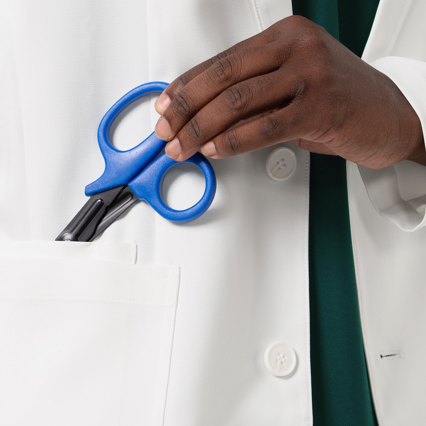 A close-up view of the LCM-201 lab coat highlights its large, practical front pocket as a hand places a pair of blue medical shears inside, emphasizing the coat’s functional storage space, clean stitching, and professional, durable white fabric designed for everyday clinical use.