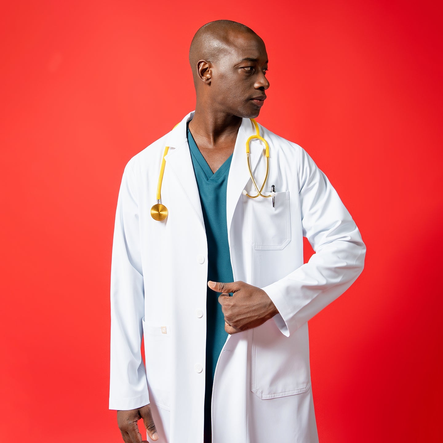 A men’s short lab coat LCM-02 in white is worn over teal scrubs, featuring a chest pocket and lower patch pockets. The model, posed against a solid red background, wears a yellow stethoscope around the neck and adjusts the front of the coat while looking to the side.