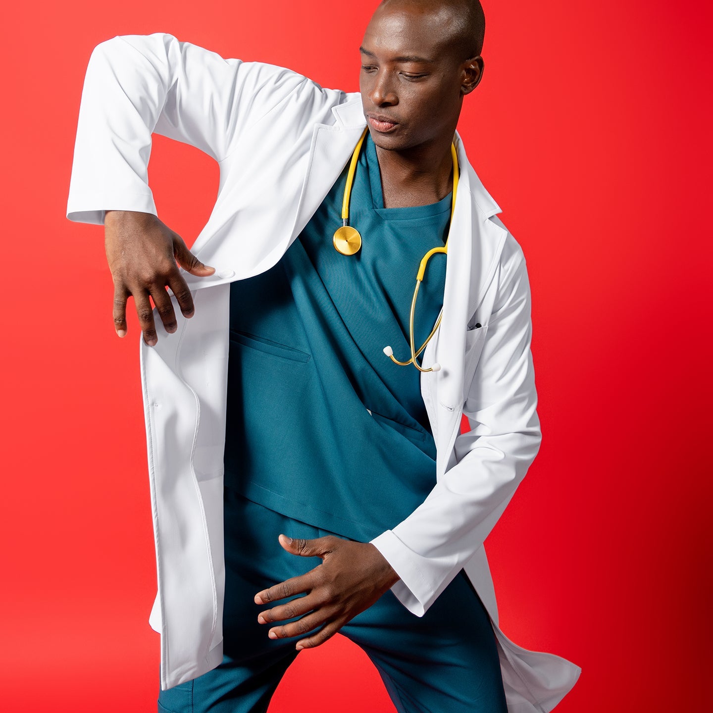 LCM-02 in white is styled over teal scrubs. The model strikes a dynamic pose against a solid red background, with a yellow stethoscope around the neck and one arm lifted to adjust the coat.