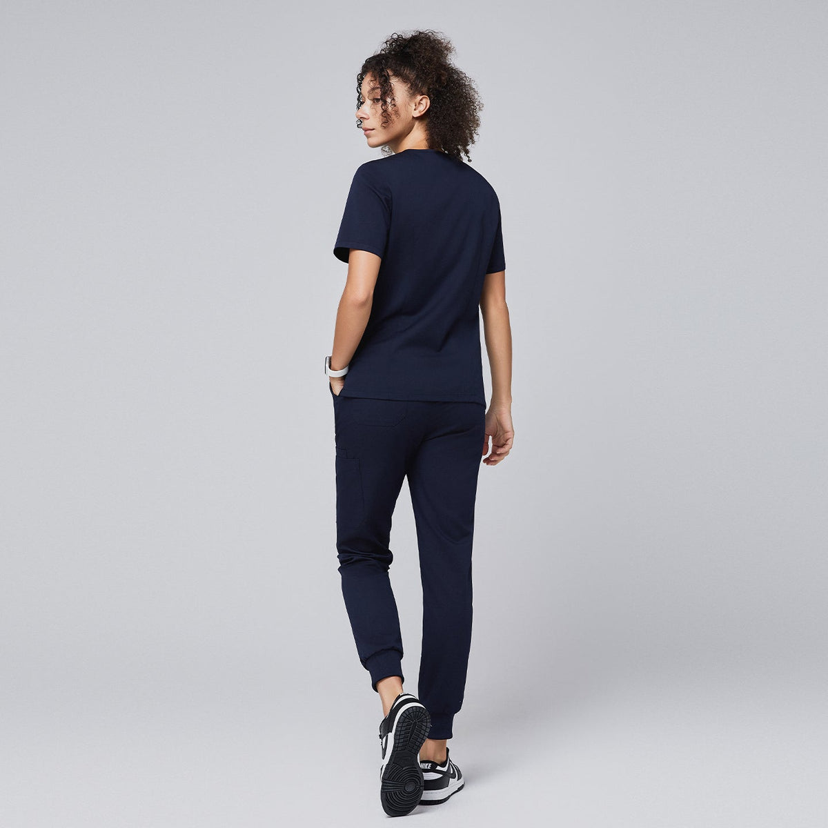 Woman wearing a navy scrub set featuring a short-sleeve top and jogger-style pants, shown from the back, with hands in pockets and black and white sneakers,Navy