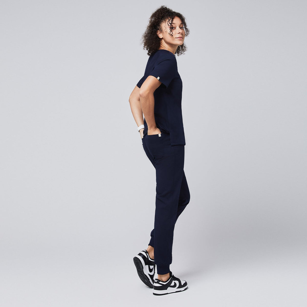 Woman wearing a navy scrub top with a chest pocket and jogger-style scrub pants, standing in a side profile pose with hands in pockets, paired with black and white sneakers,Navy