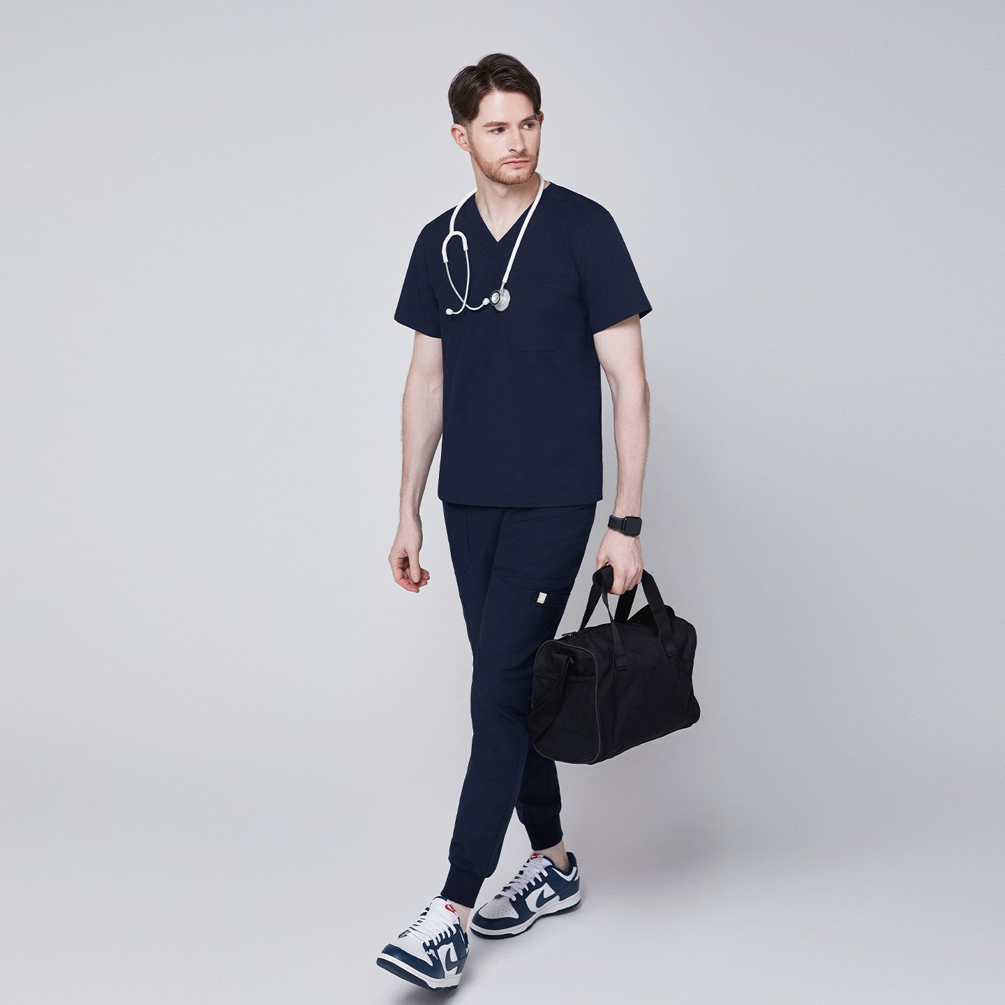 Man wearing navy V-neck scrub top and jogger scrub pants with a stethoscope and black duffle bag, paired with navy sneakers,Navy