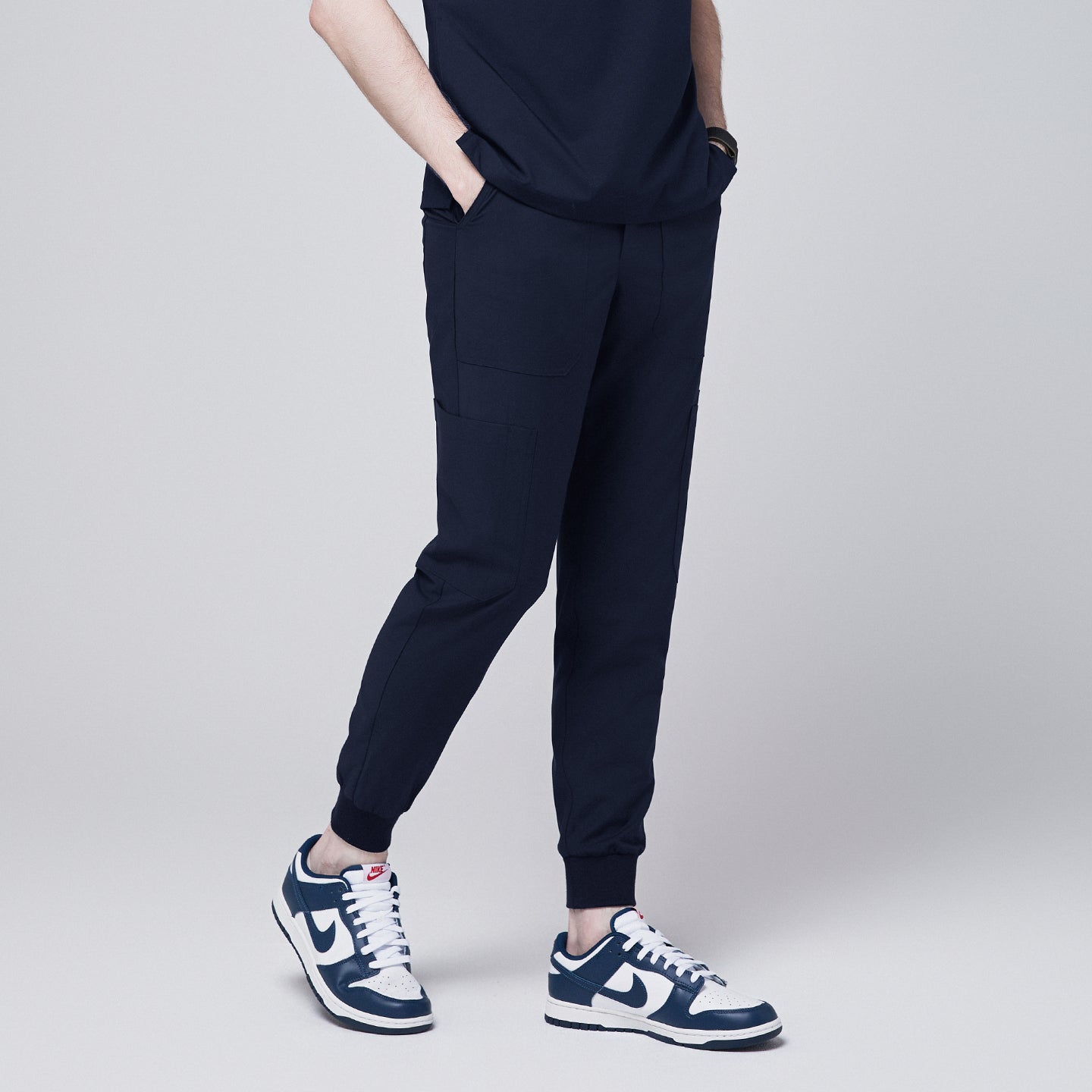 Side view of navy jogger scrub pants with 8 pockets, cuffed ankles, and hands in pockets, paired with navy sneakers,Navy