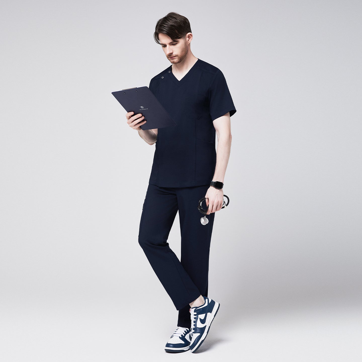 Man wearing a navy scrub set, holding a clipboard and stethoscope, showcasing the professional and modern design of the uniform, while standing in a relaxed pose,Navy