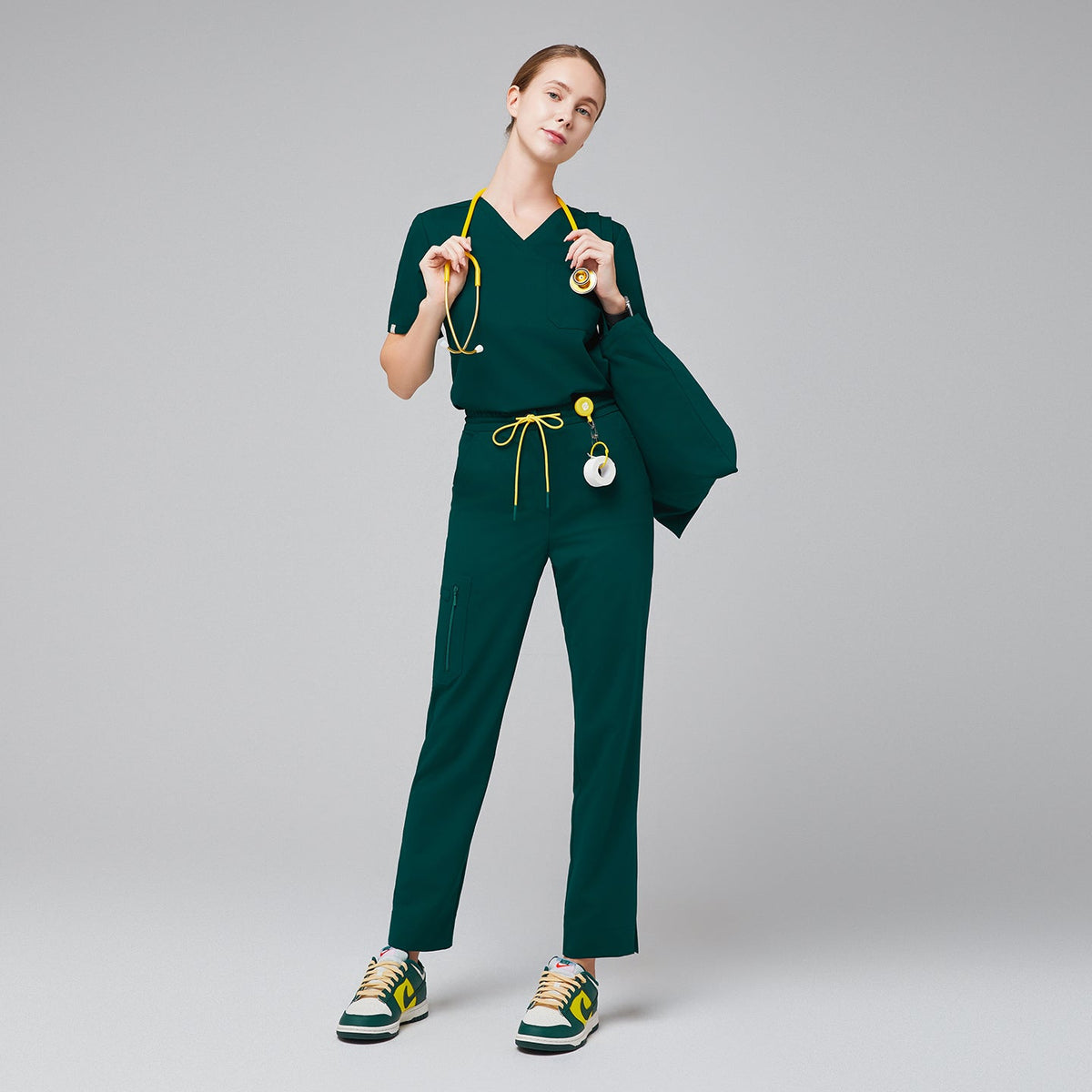 Model wearing a scrub set with a V-neck top and drawstring pants featuring zipper pockets and split hem, accessorized with a stethoscope, ID badge, and matching bag for a complete professional look,Pine Green