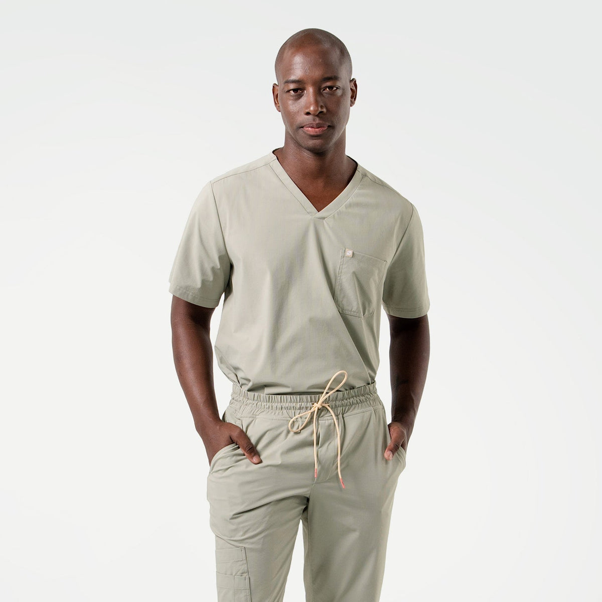The male model is shown in Sand Khaki scrubs, standing with hands in pockets and facing forward, highlighting the V-neckline and clean lines of the top paired with drawstring jogger-style pants.