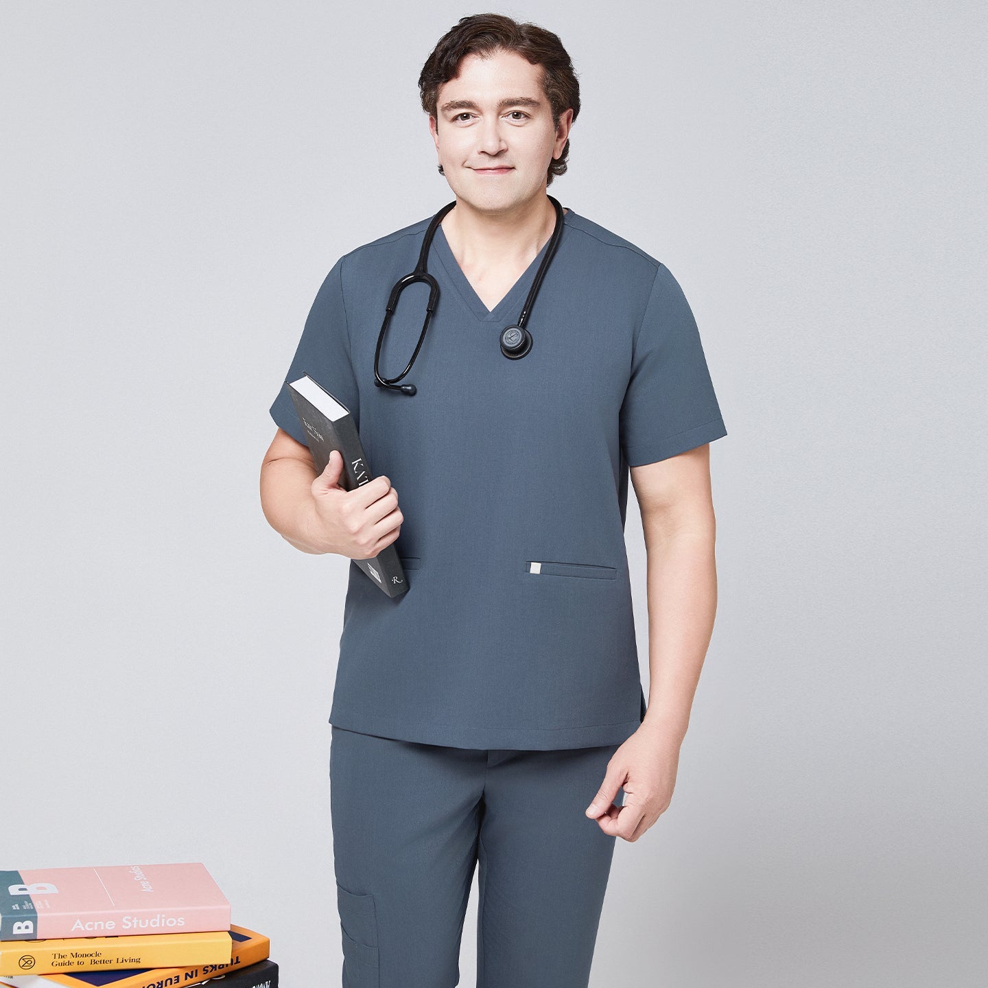 Man in a smoky blue scrub set with a double-pocket V-neck top, holding a book and wearing a black stethoscope,Smoky Blue