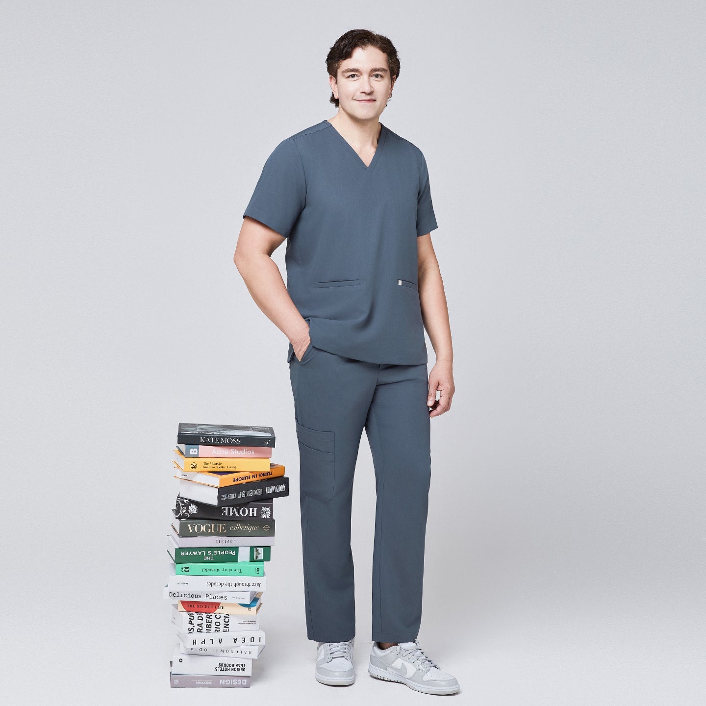 Man in a smoky blue scrub set with a double-pocket V-neck top, standing next to a stack of colorful books,Smoky Blue