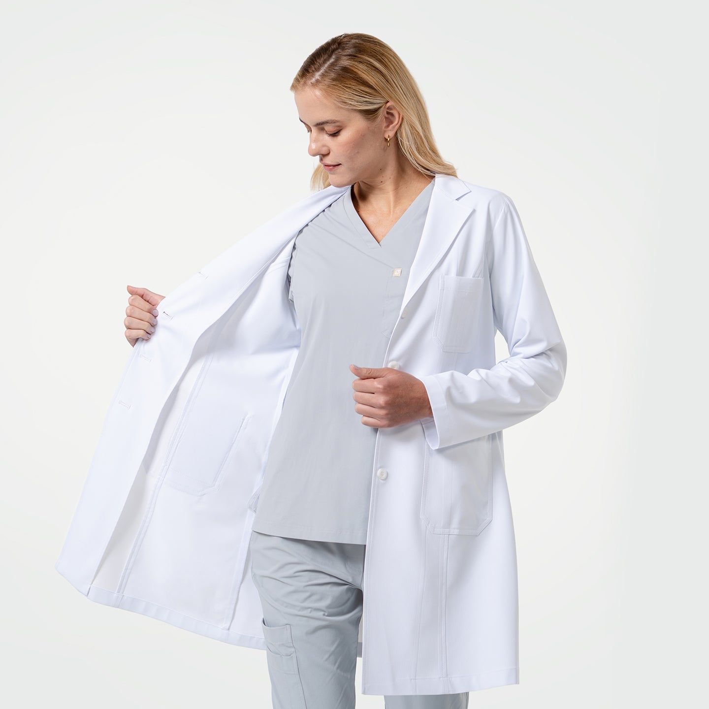 Female doctor wearing a white lab coat over light gray scrubs, looking down while opening the coat to show the inner lining on a light gray background.