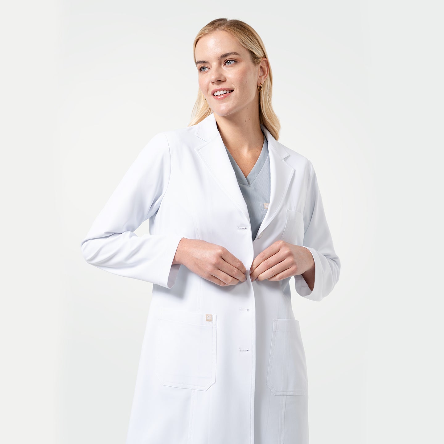 Female model fastening the front buttons of the Women’s Long Lab Coat LCW-04 in white, worn over light gray scrubs.
