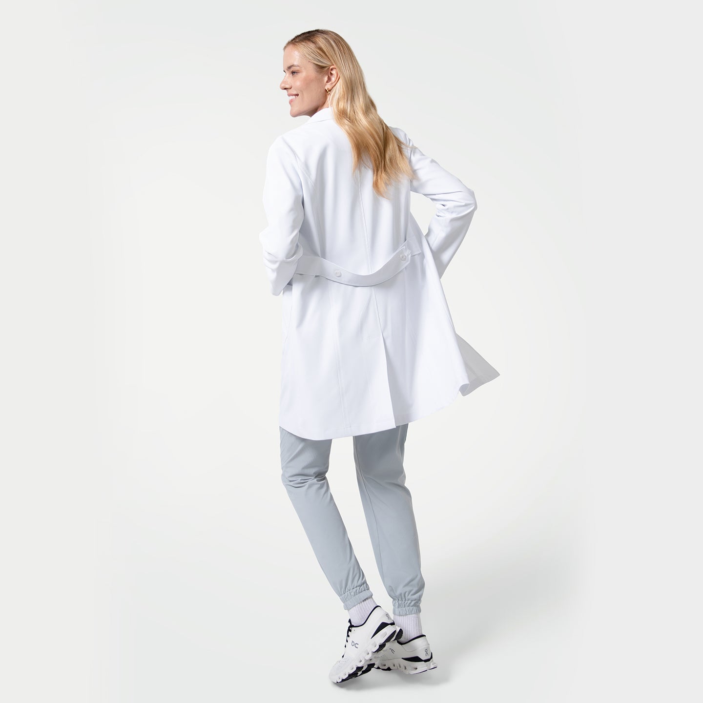 Model facing away from the camera wearing the Women’s Long Lab Coat LCW-04 in white, showing the back belt detail, paired with light gray jogger scrub pants and white sneakers.