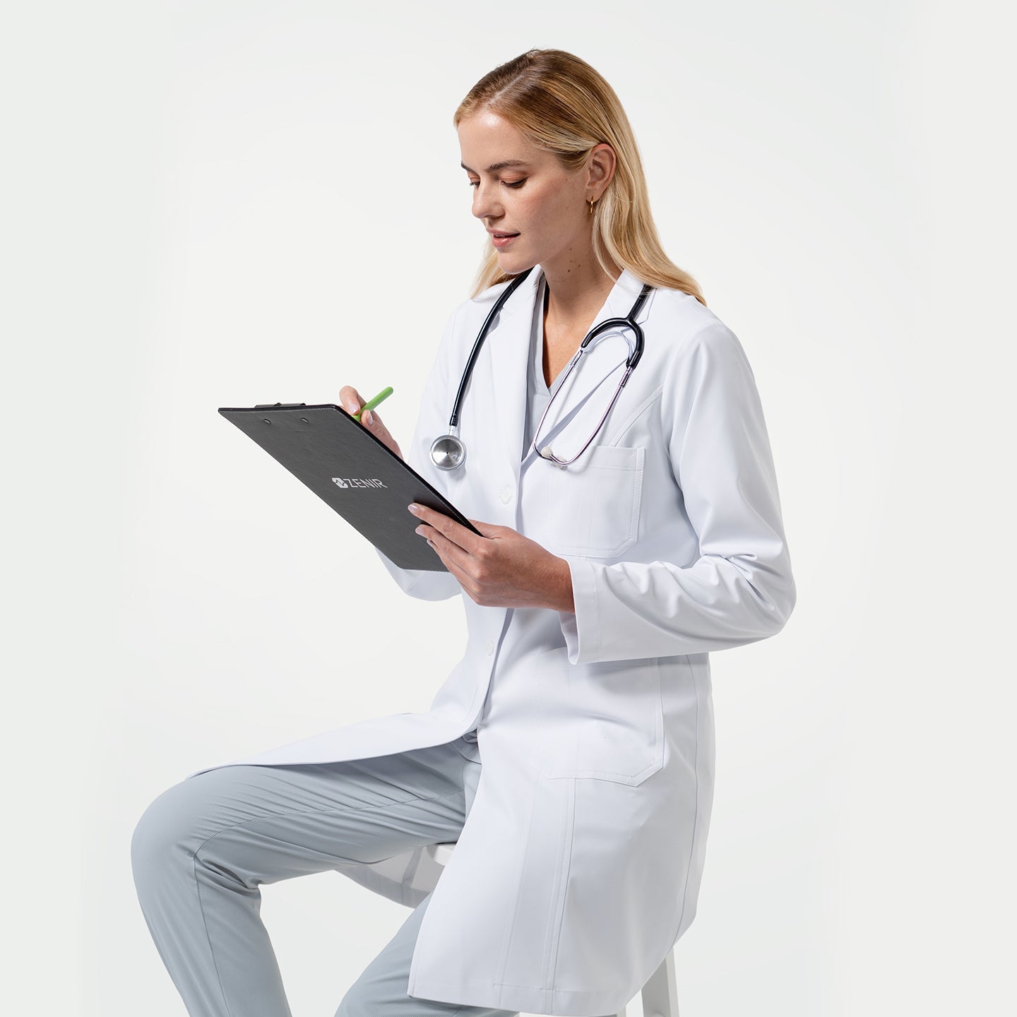 Model wearing a white medical lab coat and stethoscope, sitting on a stool while taking notes on a clipboard against a light gray background.