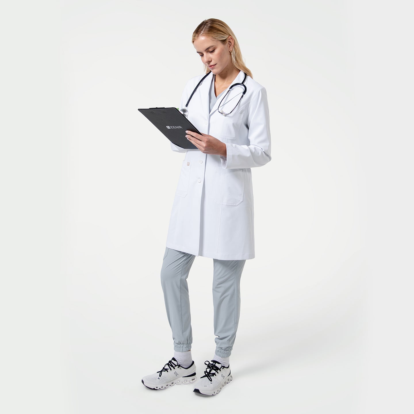 Female doctor wearing a white lab coat with a stethoscope around her neck, holding a clipboard and reviewing notes on a light gray background.