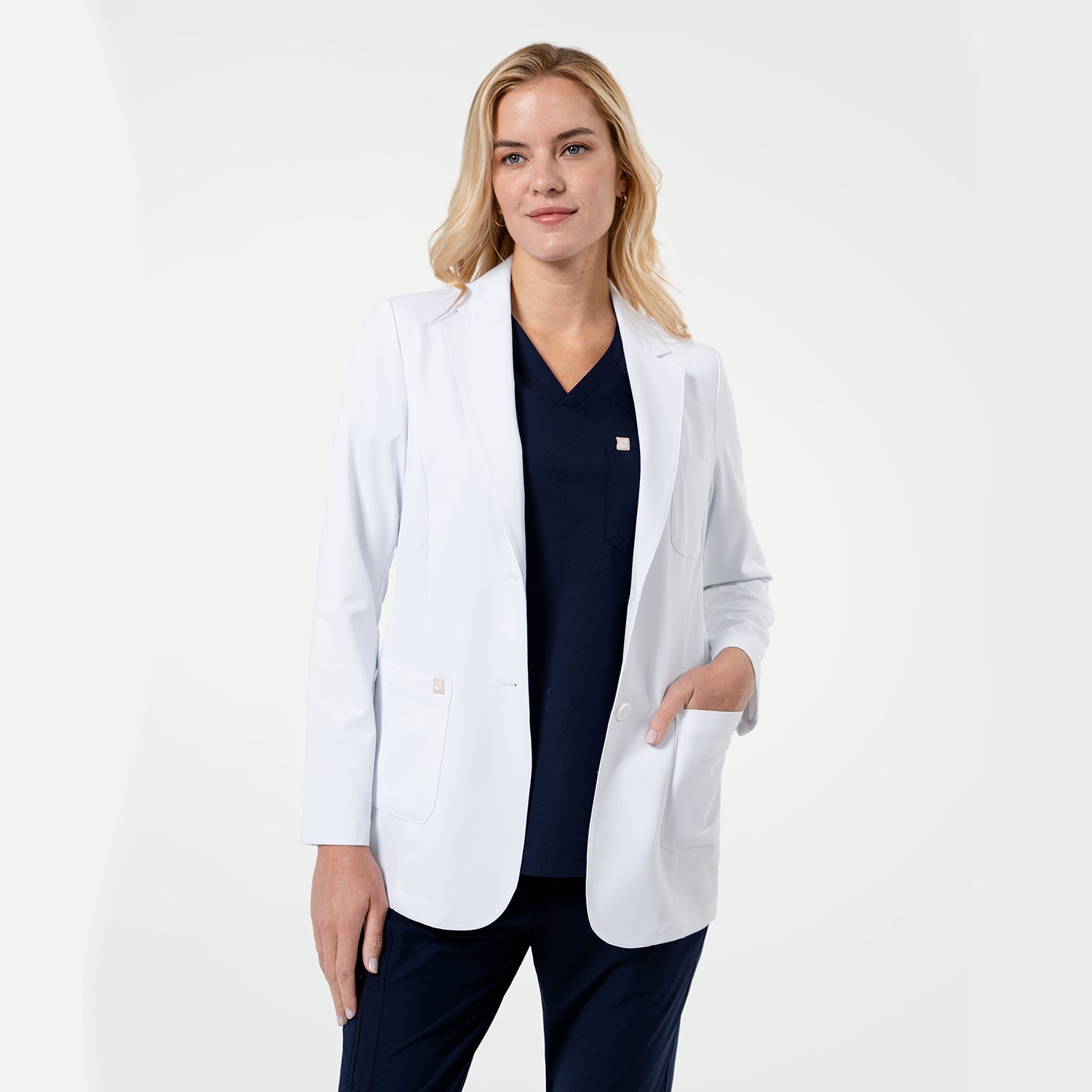 Confident female healthcare professional wearing a white coat layered over navy scrubs, standing with one hand in her pocket on a clean light gray background.