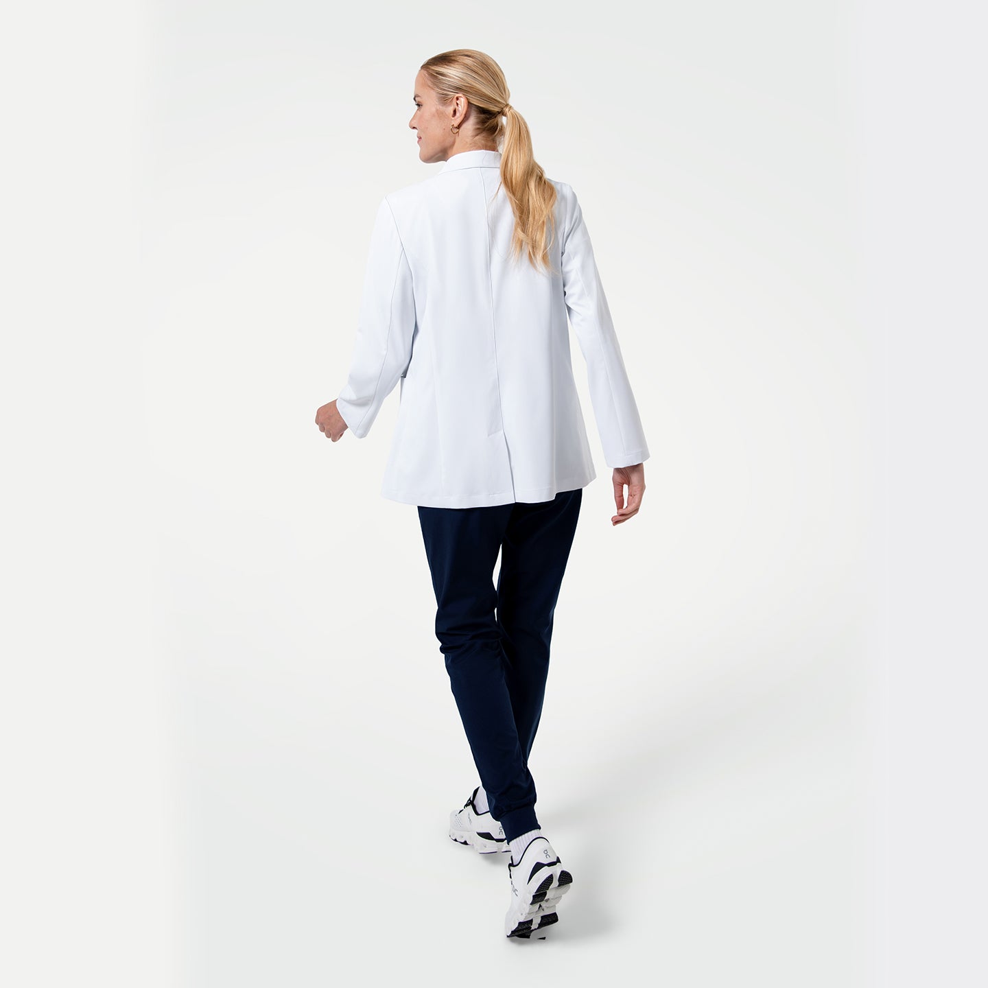 Back view of the Women’s Short Lab Coat LCW-02 in white, showing the model walking away with the coat’s straight hemline and side vents visible, paired with navy scrub pants and white sneakers.