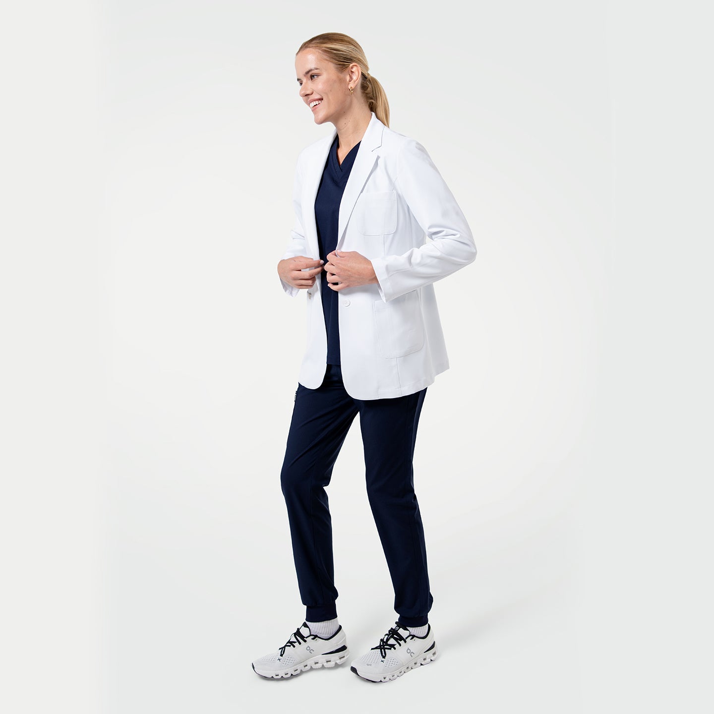Model wearing a white medical lab coat layered over navy scrubs, smiling and adjusting the front of the coat on a light gray background.