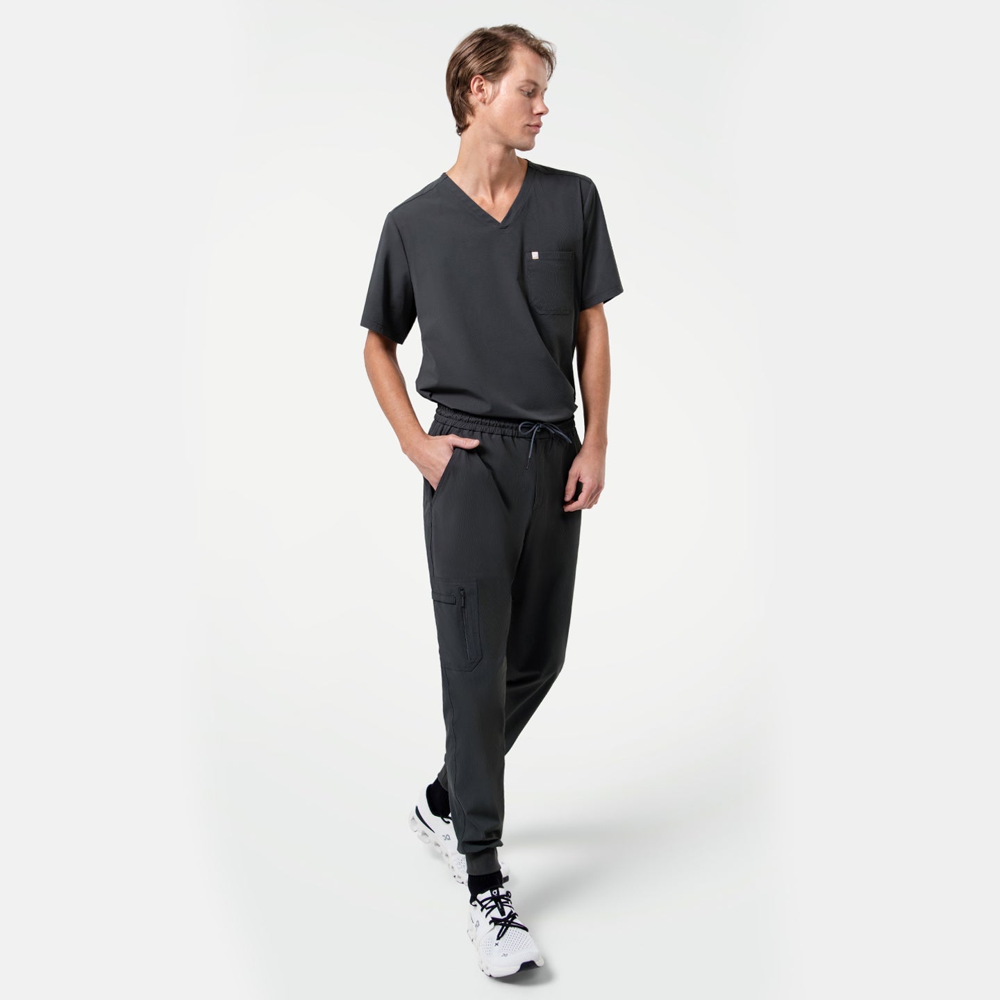 Male model walking with hands in pockets, wearing a matching V-neck scrub top and Zipper Pocket Jogger Scrub Pants in Deep Gray, paired with white sneakers.