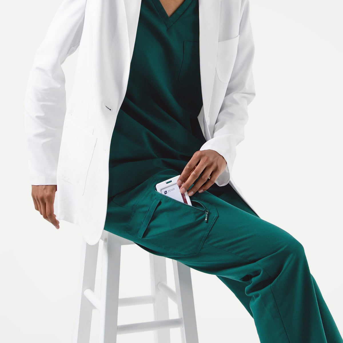 Person wearing a white lab coat over Pine Green zipper pocket scrub pants, seated on a white stool while placing a phone into the pants pocket.