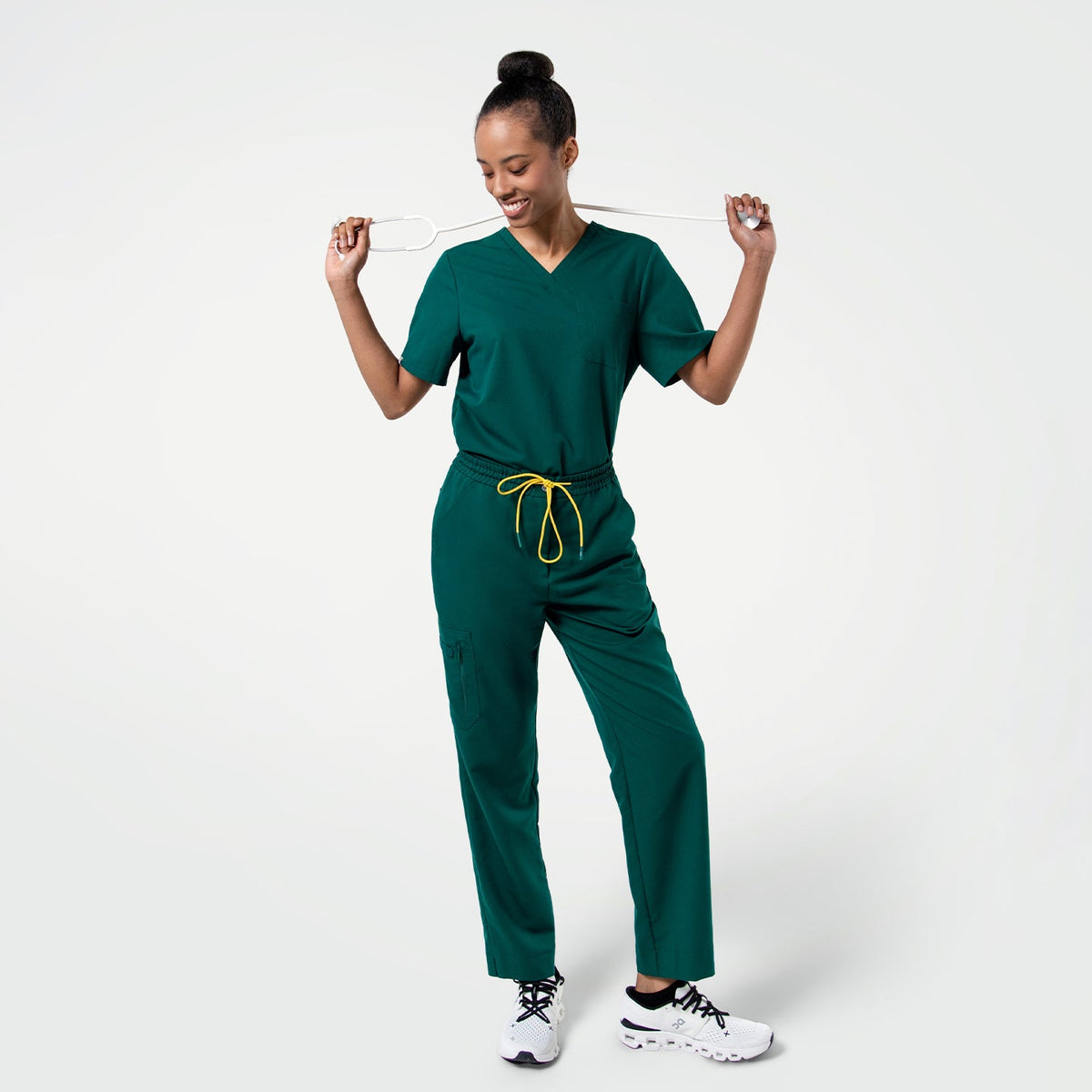 Person wearing Pine Green zipper pocket split hem scrub pants and a matching V-neck scrub top, standing with a stethoscope held playfully across their shoulders.